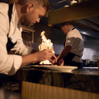 man plating food and fire behind him in a kitchen